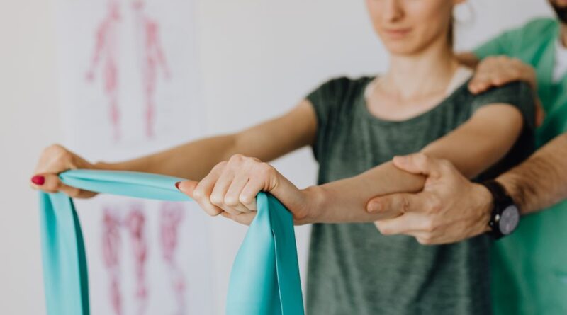 Crop anonymous orthopedist in wristwatch and uniform helping young woman in casual wear reaching arms with elastic tape in doctor office - Photo by https://kaboompics.com/ on Pexels