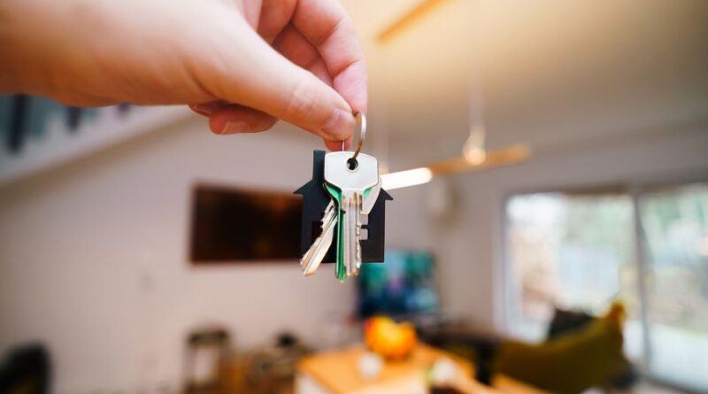 A hand holds keys with a house keychain inside a modern, well-lit living space. - Photo by Jakub Zerdzicki on Pexels